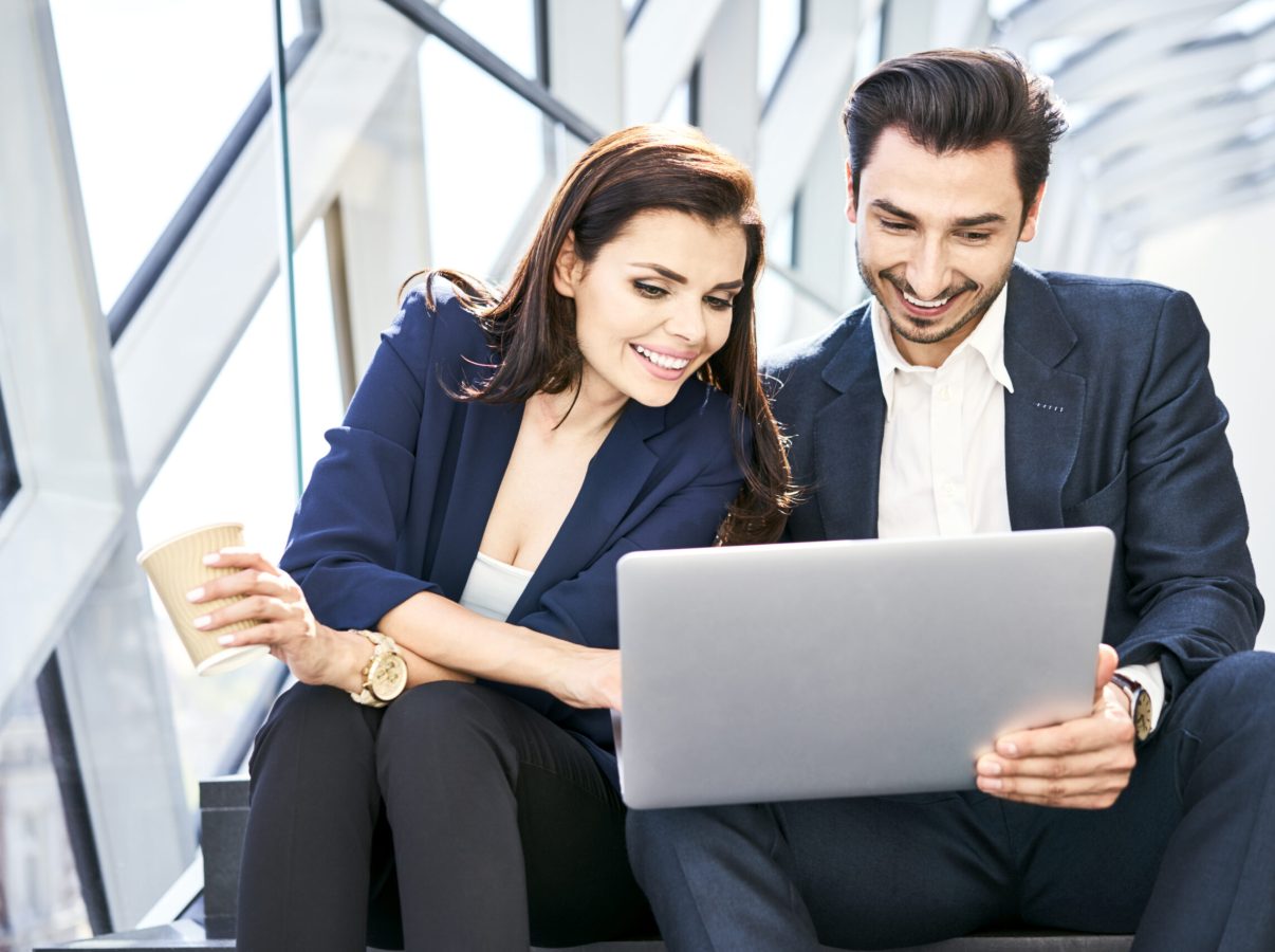 Smiling businesswoman and businessman sharing laptop on stairs in modern office