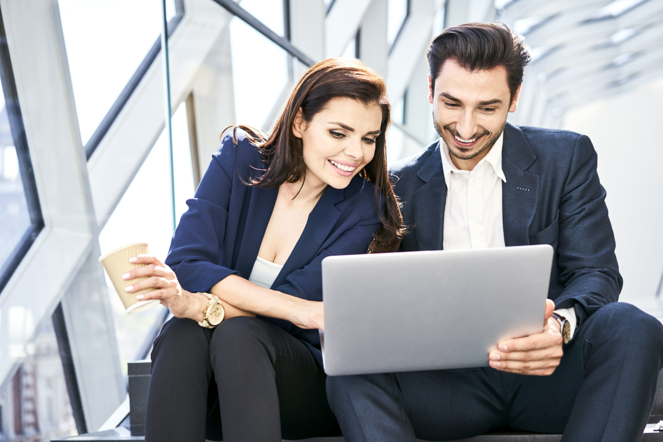 Smiling businesswoman and businessman sharing laptop on stairs in modern office